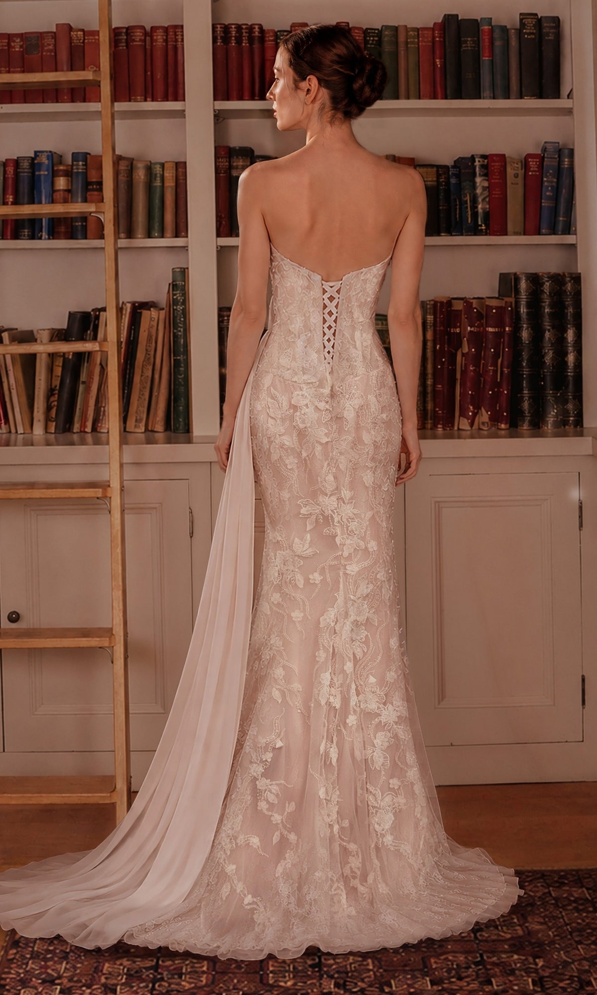 A woman in the Ladivine CB168W Long Prom Dress, a strapless gown adorned with delicate floral lace appliques and featuring a sheer train, stands in front of a bookcase, facing away from the camera.